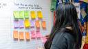 Listening session participant looking at a wall with sticky notes holding a pink sticky note with the words road, project, updates written on it. Listening session participant looking at a wall with sticky notes holding a pink sticky note with the words road, project, updates written on it.
