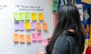 Listening session participant looking at a wall with sticky notes holding a pink sticky note with the words road, project, updates written on it.